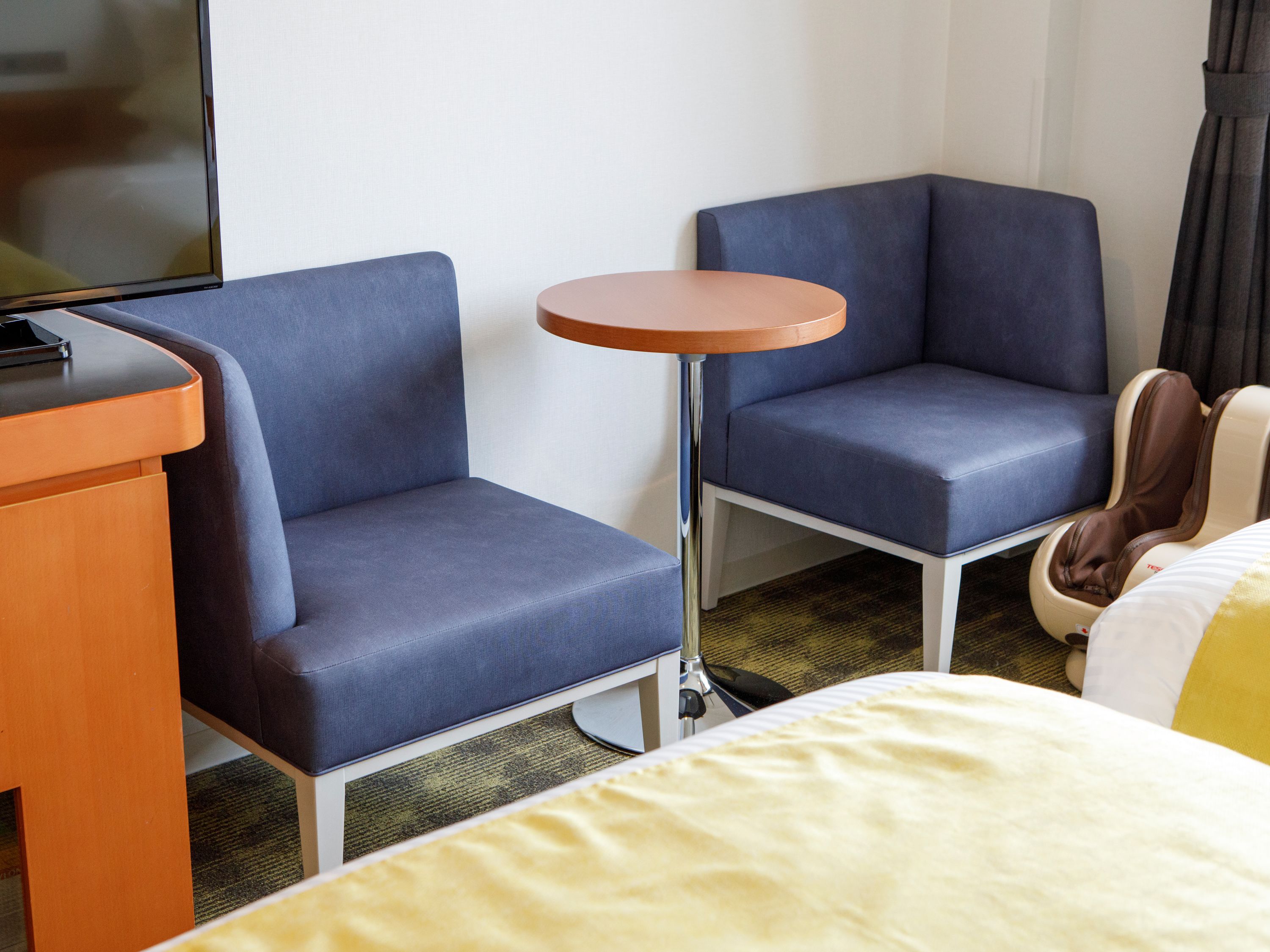 A hotel room with two blue armchairs, a small round table, a bed with a yellow bedspread, and a television on a wooden desk.