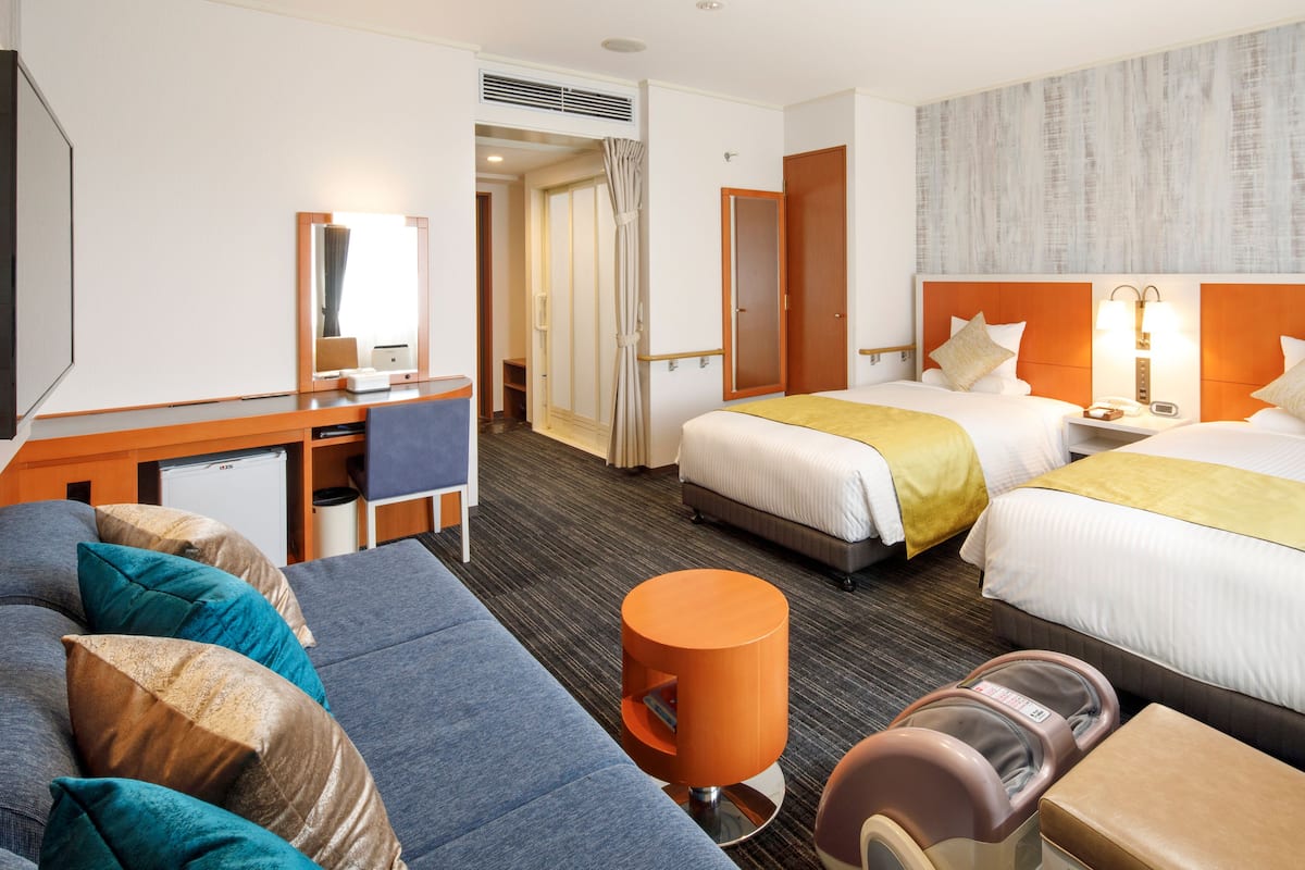 A hotel room with a sofa, two beds, a desk, a chair, a massage chair, and a small orange table.