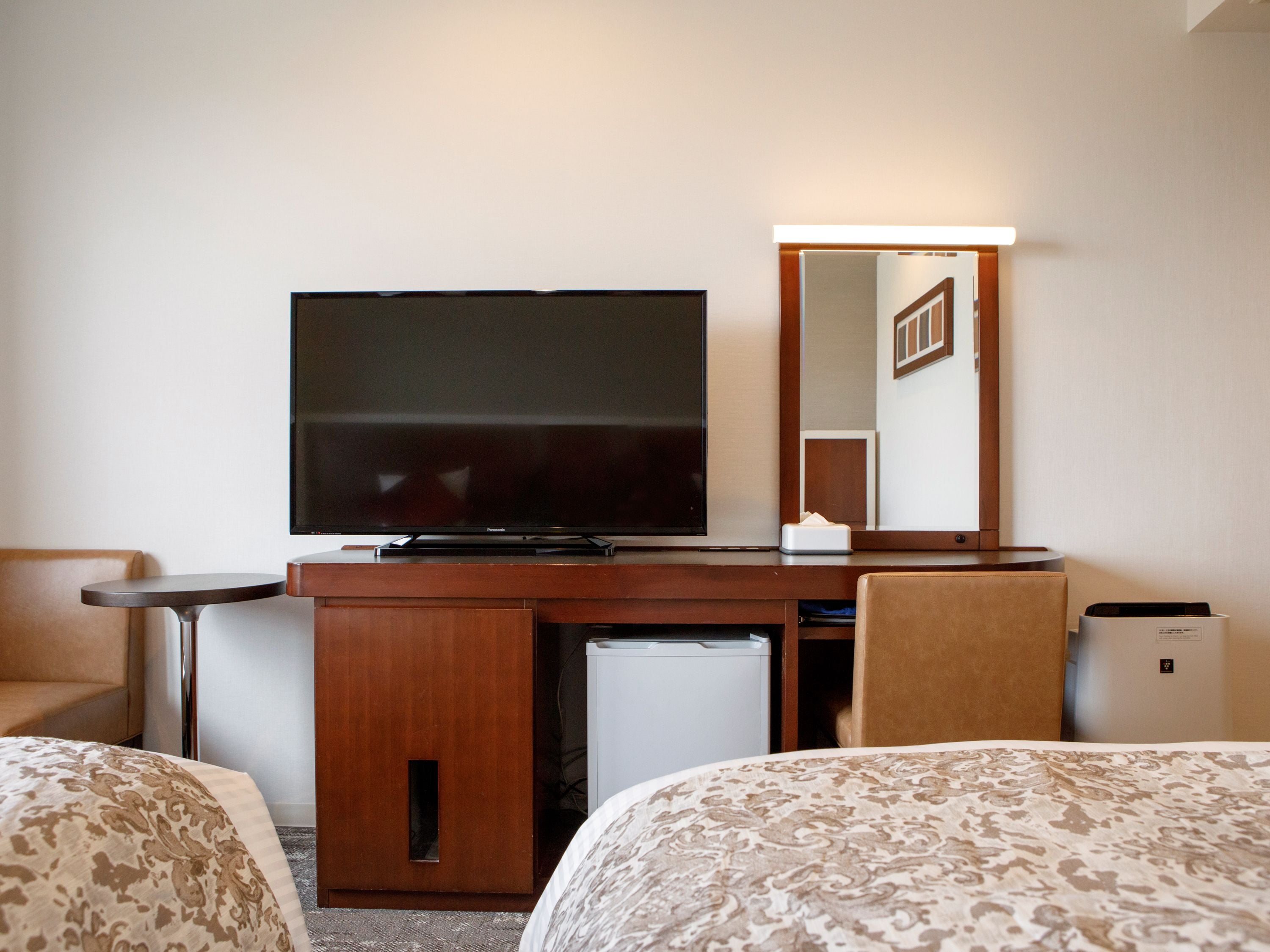 Habitación de hotel con televisión de pantalla plana, escritorio con lámpara, refrigerador pequeño, espejo y dos camas con ropa de cama estampada.