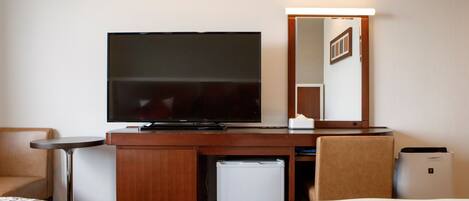 Habitación de hotel con televisión de pantalla plana, escritorio con lámpara, refrigerador pequeño, espejo y dos camas con ropa de cama estampada.