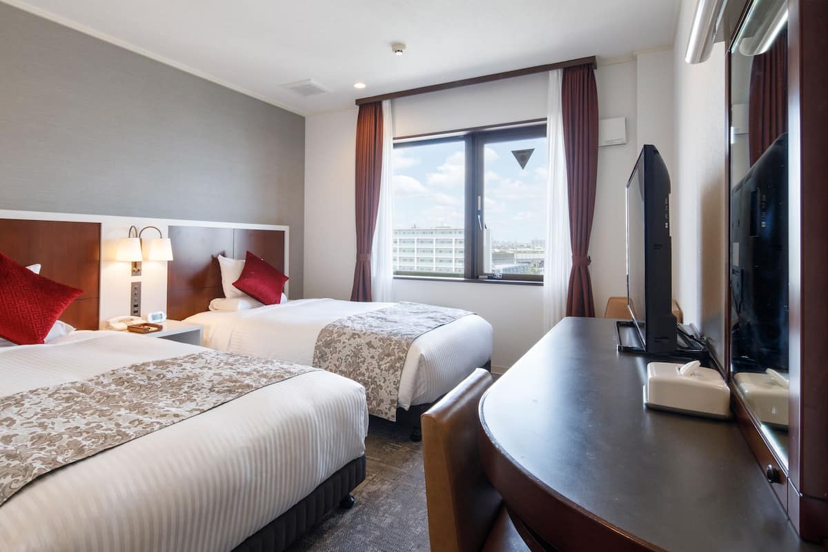 A hotel room with two beds, a desk, a chair, a TV, and a window with curtains.