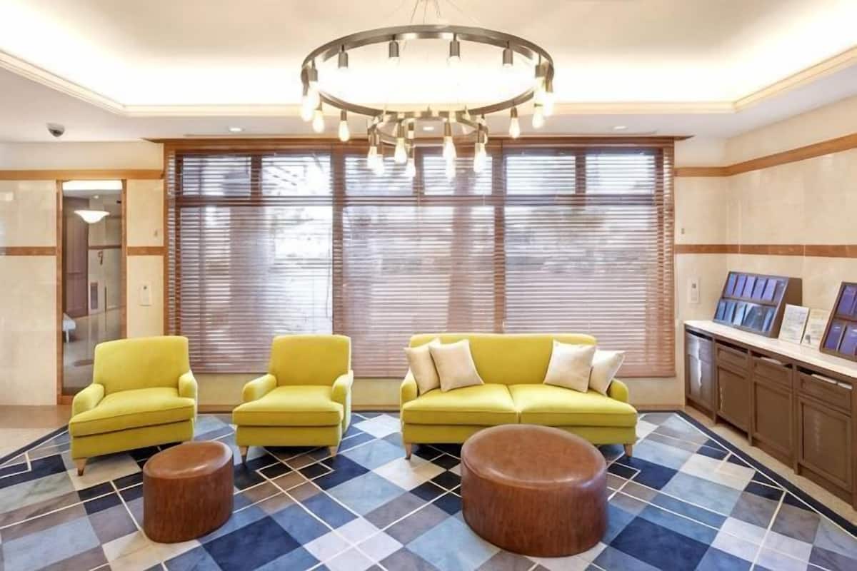 A lobby with three yellow armchairs, a blue and white checkered floor, a chandelier, and large windows with blinds.