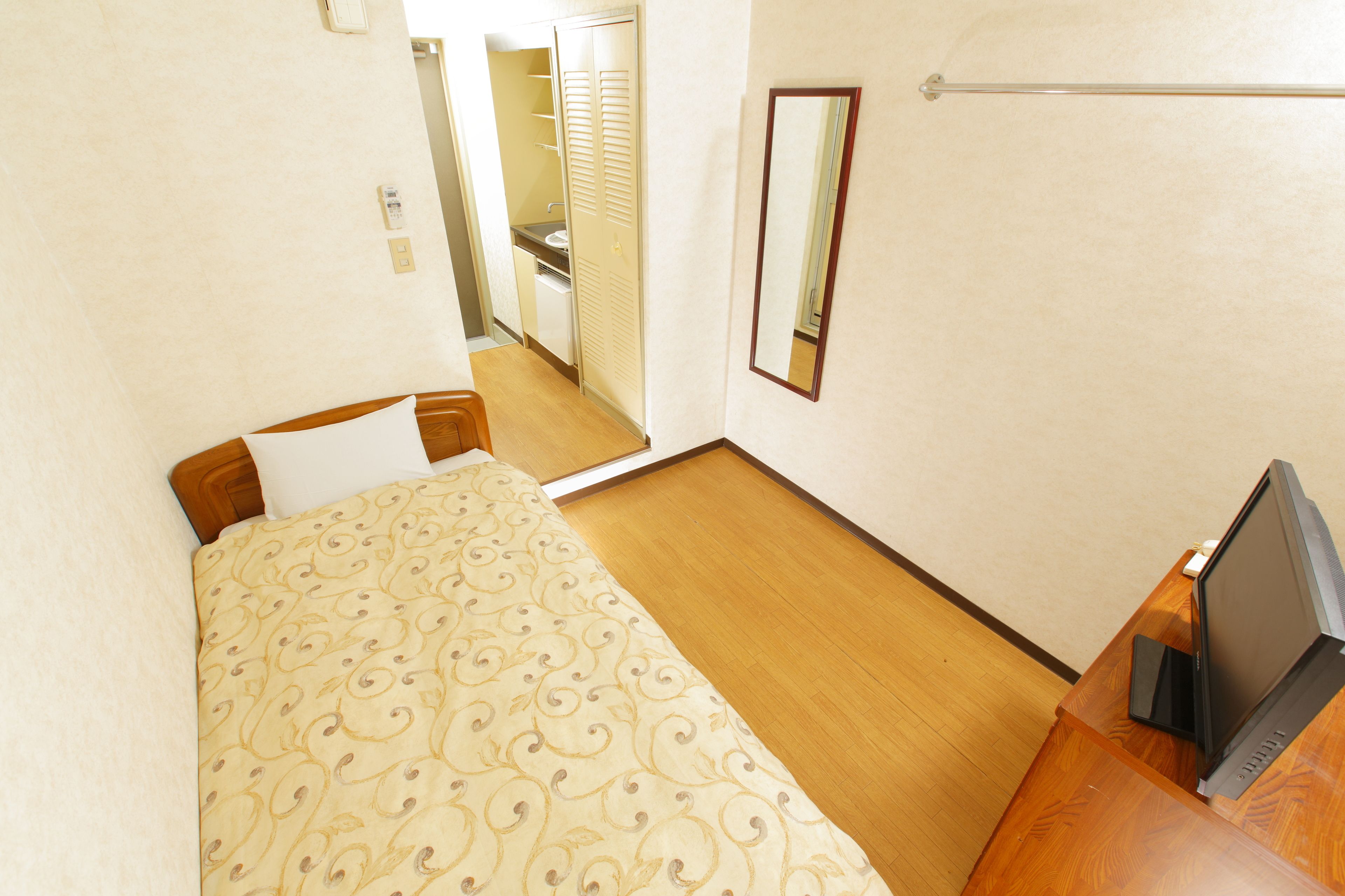Un dormitorio con cama, un escritorio de madera con una computadora portátil, un espejo y un baño visible a través de una puerta abierta.