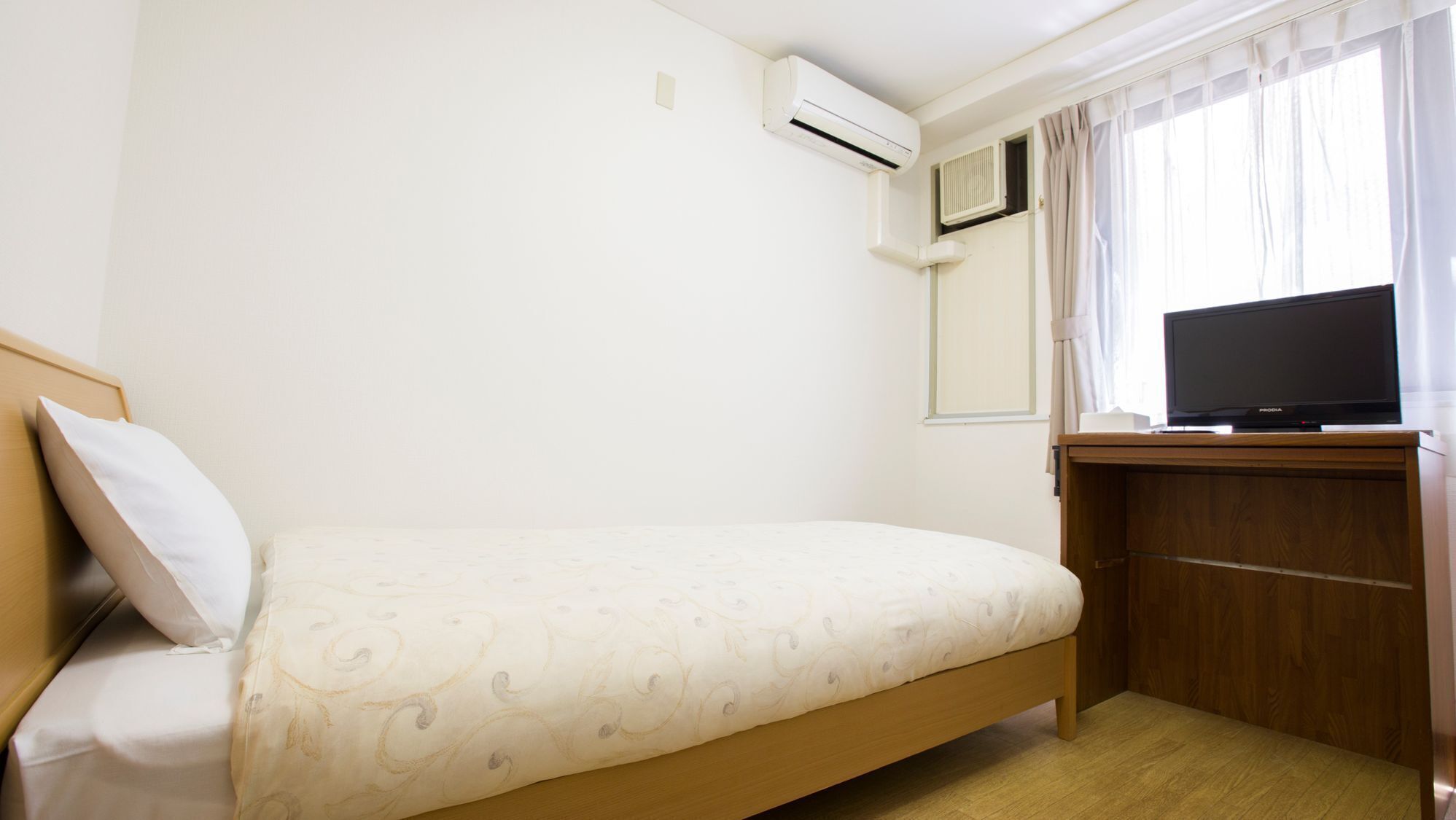 A hotel room with a bed, an air conditioning unit, a television on a wooden stand, and a window with curtains.