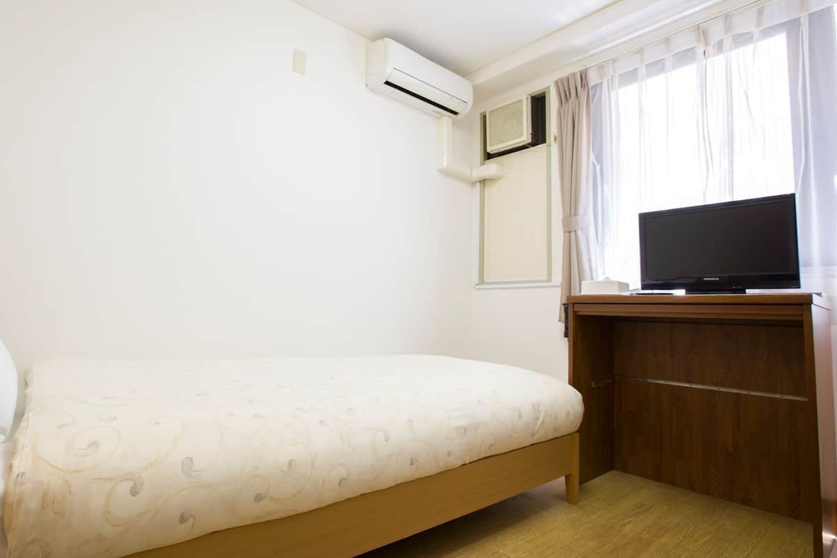 Una habitación de hotel con una cama, un equipo de aire acondicionado, un televisor sobre un soporte de madera y una ventana con cortinas.