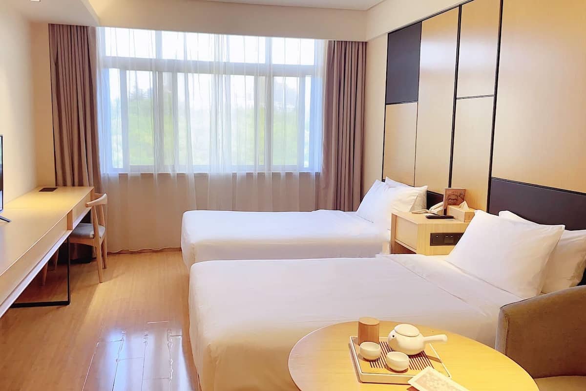 Une chambre d’hôtel avec deux lits, un bureau avec une télévision, une table de chevet avec un téléphone et un livre, et une fenêtre avec des rideaux.