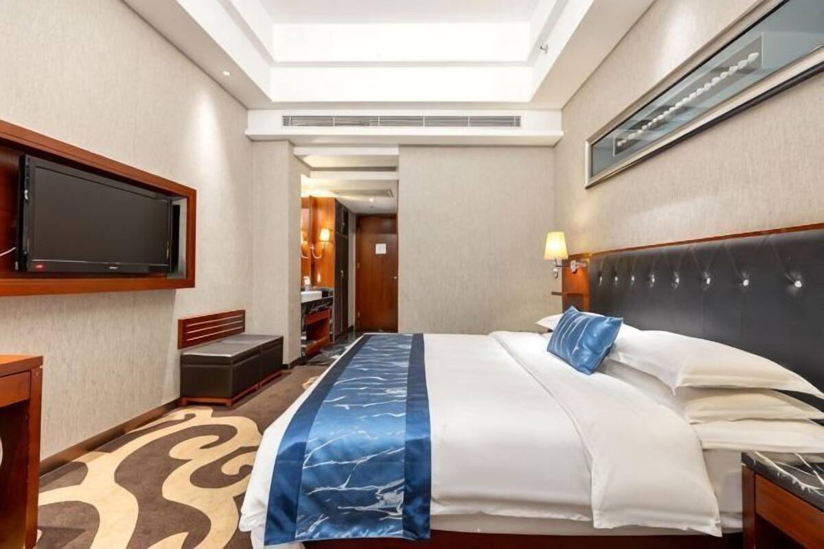 A hotel room with a bed, a TV, a headboard, a lamp, and a patterned carpet.