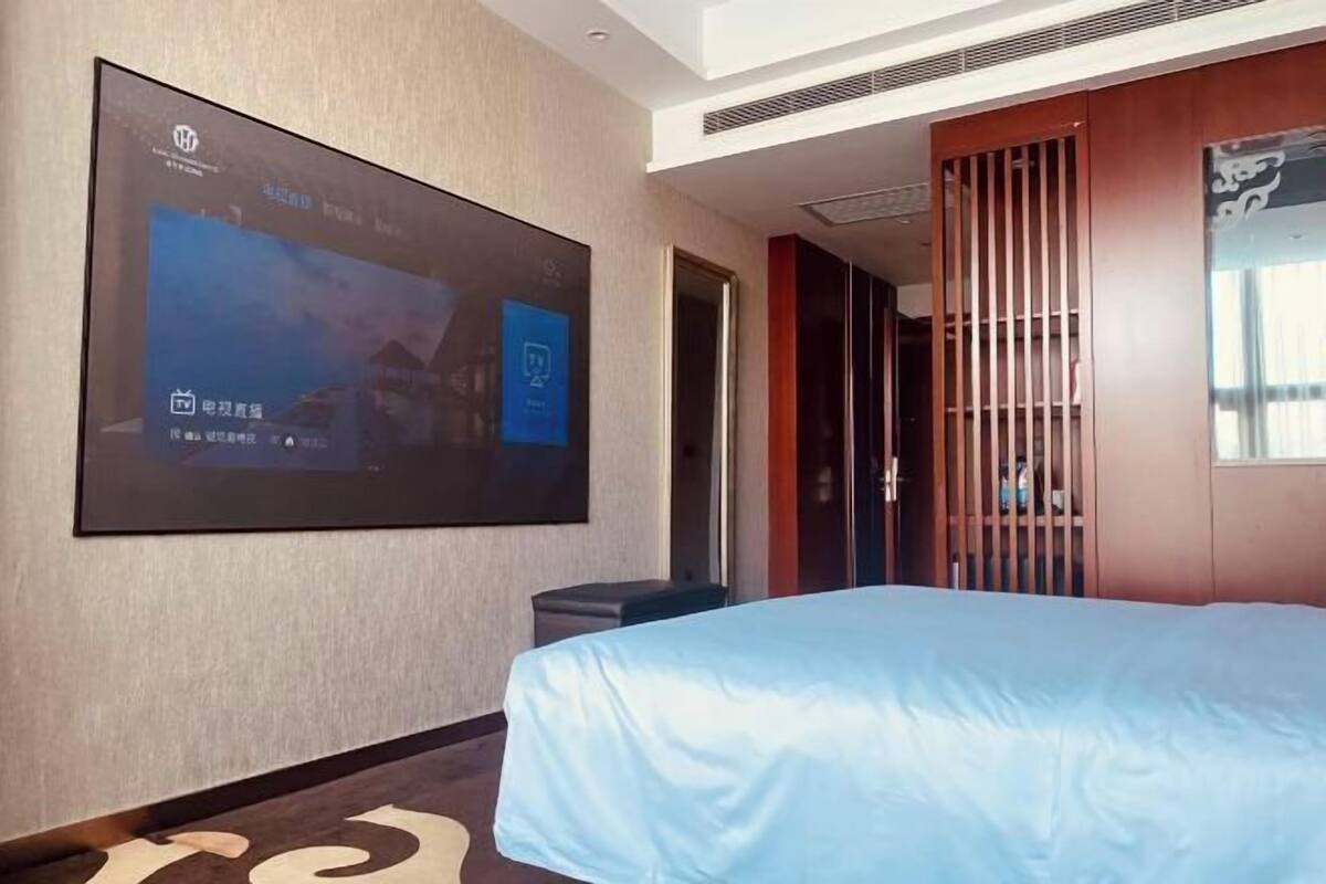A hotel room with a large TV mounted on the wall, a bed with a blue cover, a wooden wardrobe, and a door leading to another room.