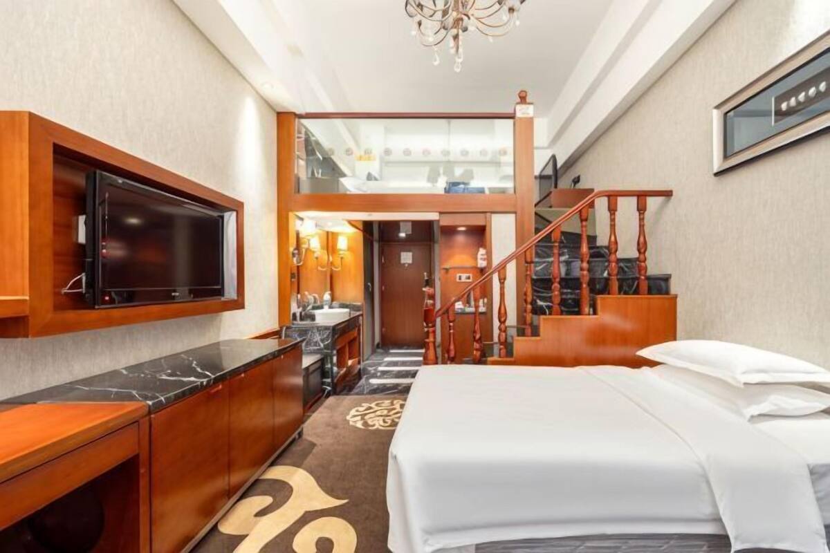 A hotel room with a bed, a TV mounted on the wall, a wooden staircase, and a bathroom visible through an open door.