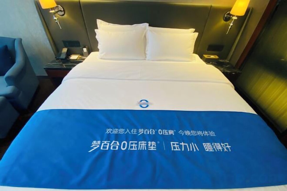 A hotel room with a neatly made bed, a blue bedspread with text, a nightstand with a lamp, and a chair.