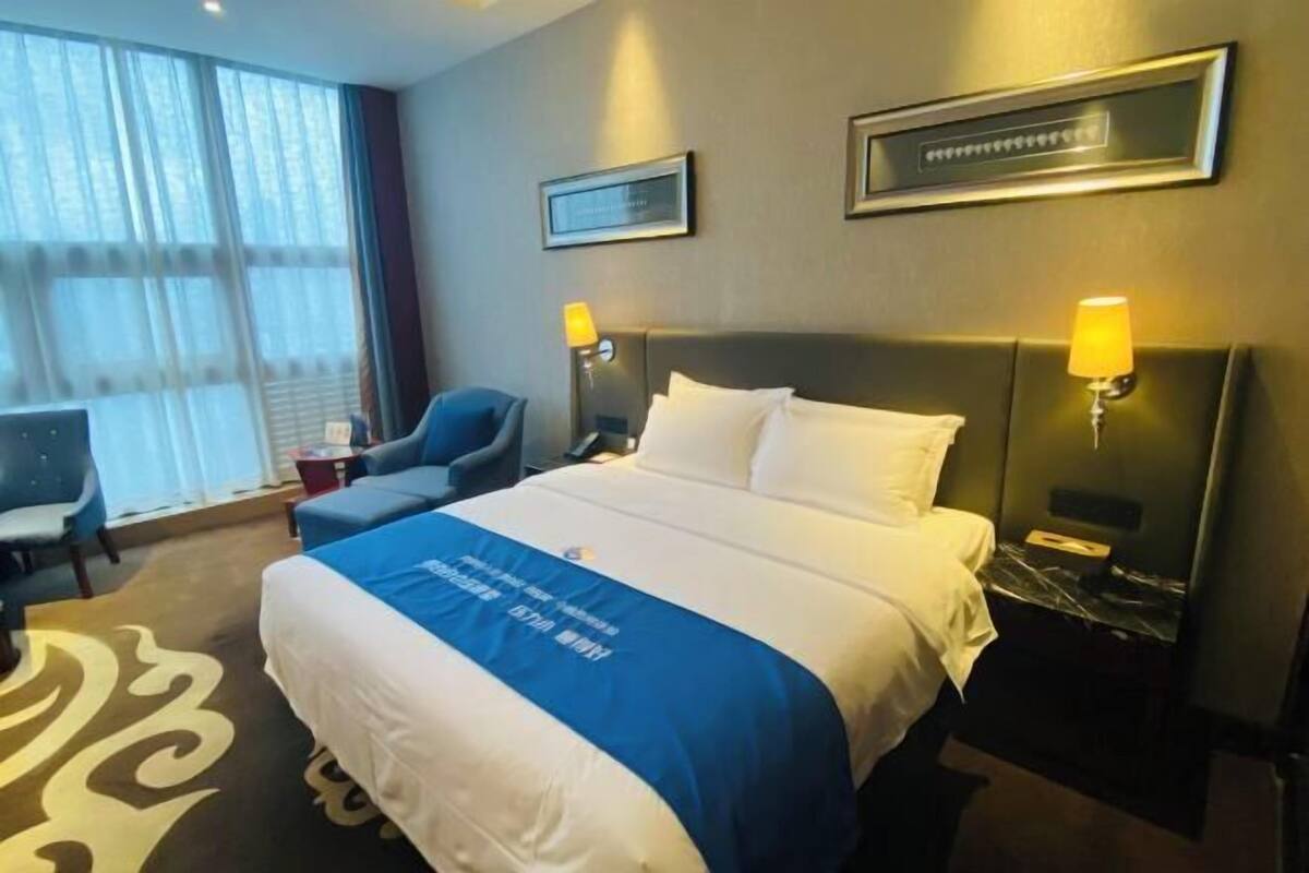 A hotel room with a bed, two bedside lamps, a chair, a desk, and a large window with curtains.