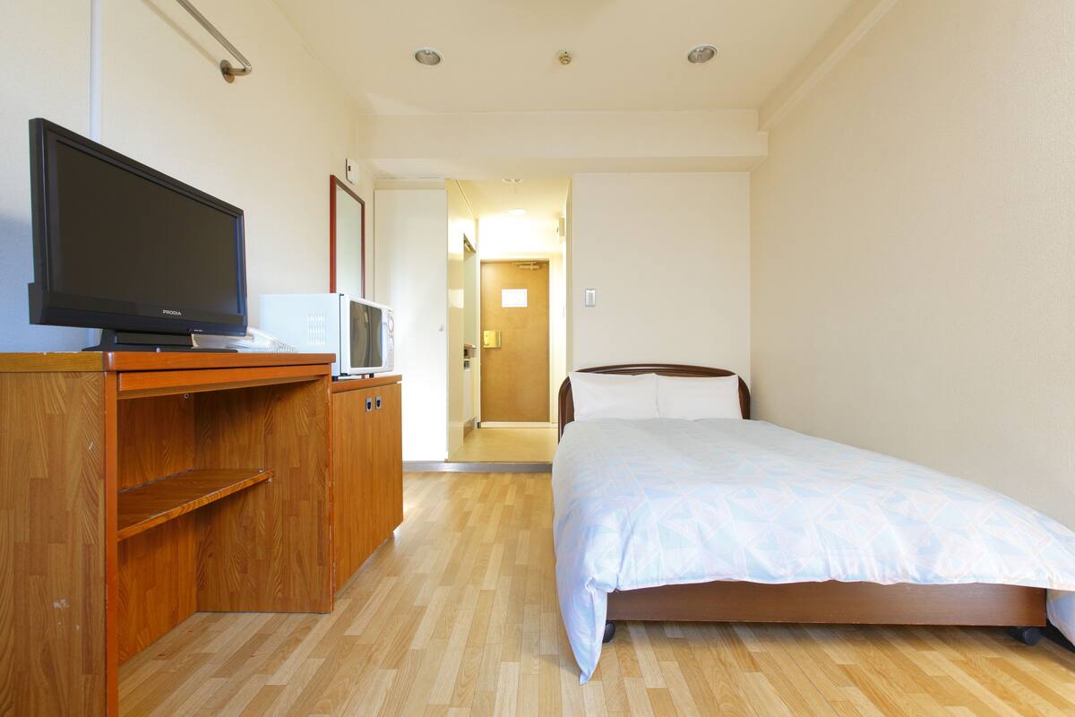 A hotel room with a wooden desk, a television, a bed with a white bedspread, and a wooden floor.