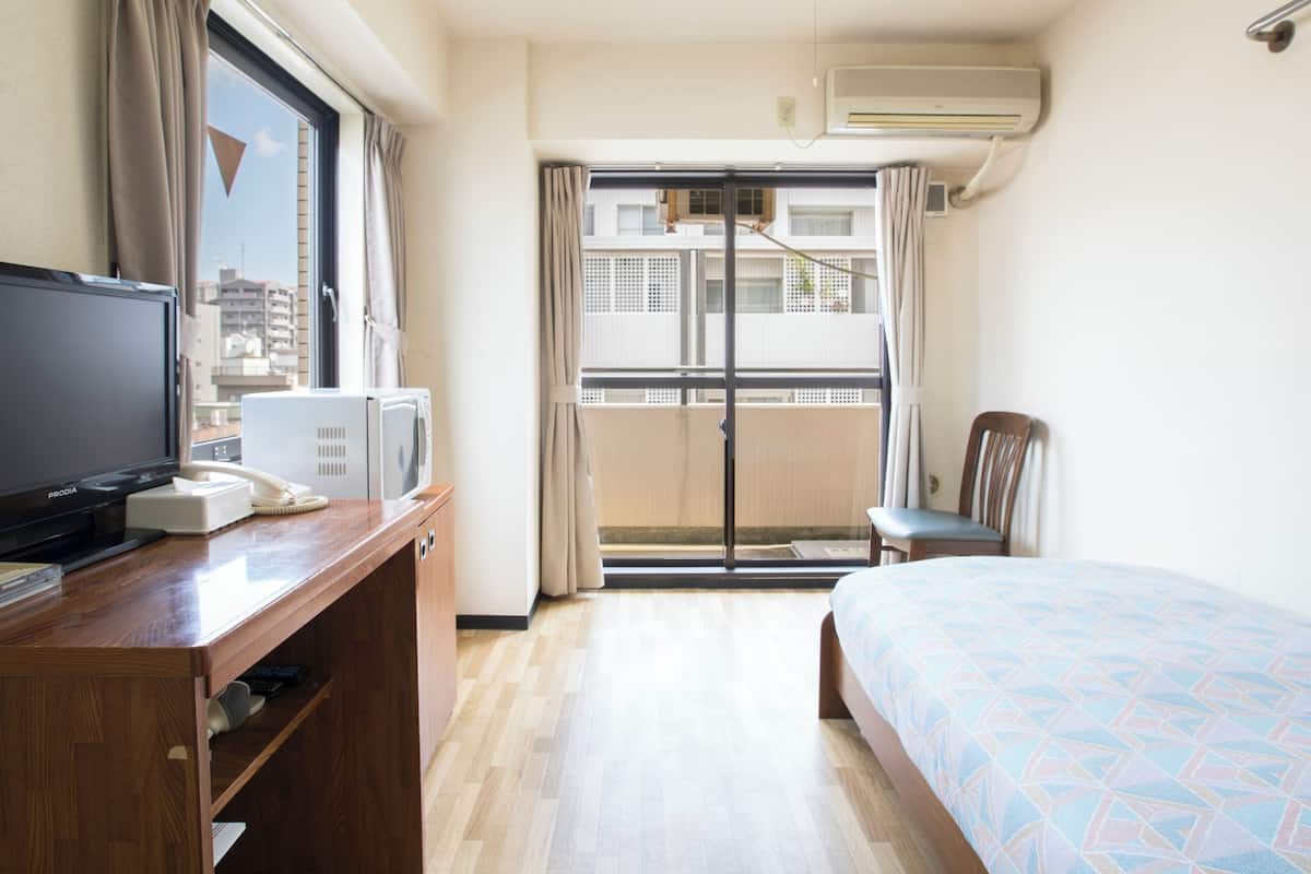 A hotel room with a bed, a desk with a TV, a phone, and a microwave. There is a chair and a window with curtains.