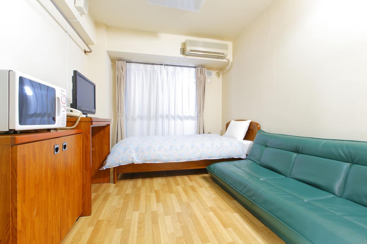A compact room with a bed, a green sofa, a wooden cabinet, a television, and an air conditioning unit.