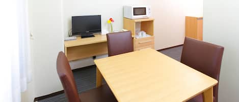 A hotel room with a wooden table, four brown chairs, a television, a microwave, and a small cabinet with a vase of flowers.