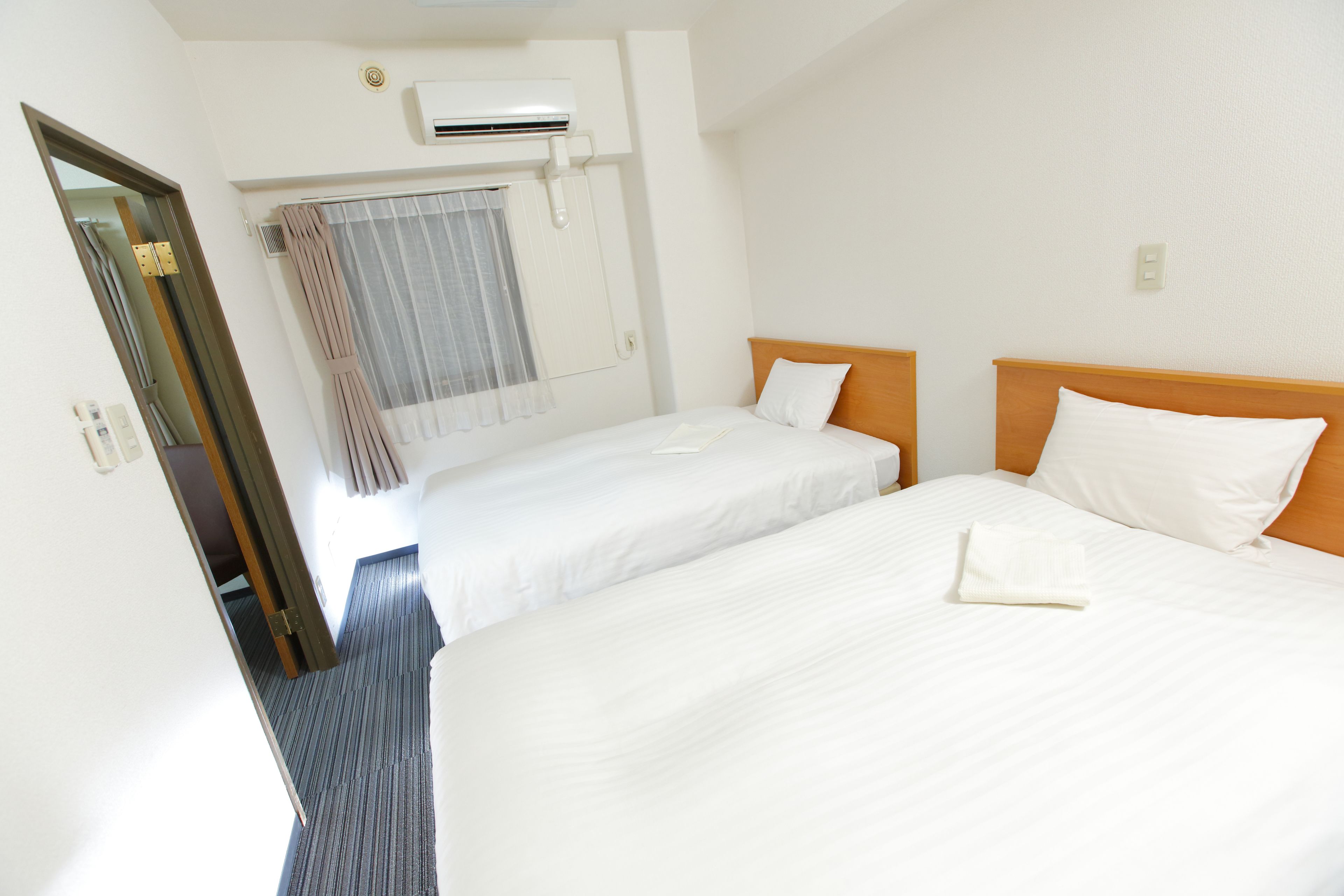 Una habitación de hotel con dos camas individuales, una ventana con cortinas, un equipo de aire acondicionado y una puerta que da a otra habitación.
