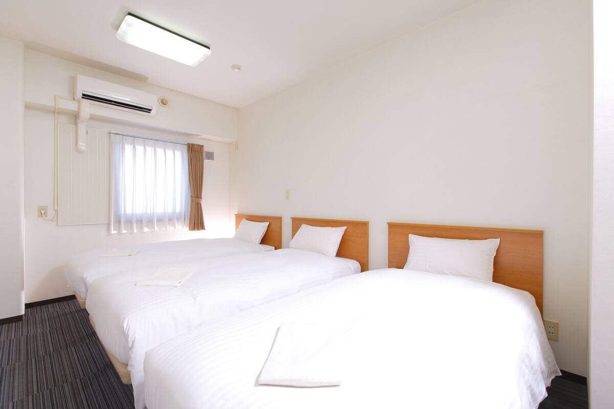 Une chambre d’hôtel avec deux lits simples, une grande fenêtre avec des rideaux, un appareil de climatisation et un sol recouvert de moquette.