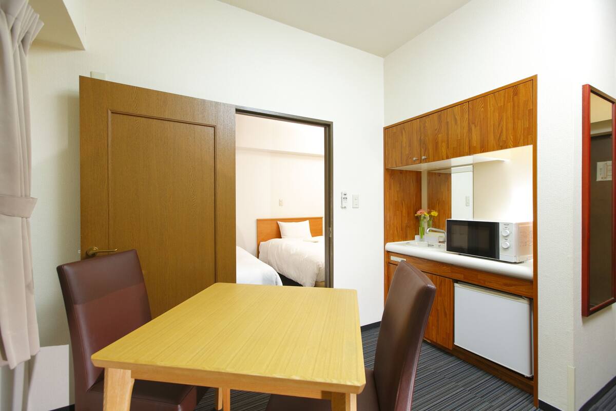 Une chambre d’hôtel avec une table en bois, deux chaises, un lit avec une literie blanche, un four à micro-ondes et un petit réfrigérateur.