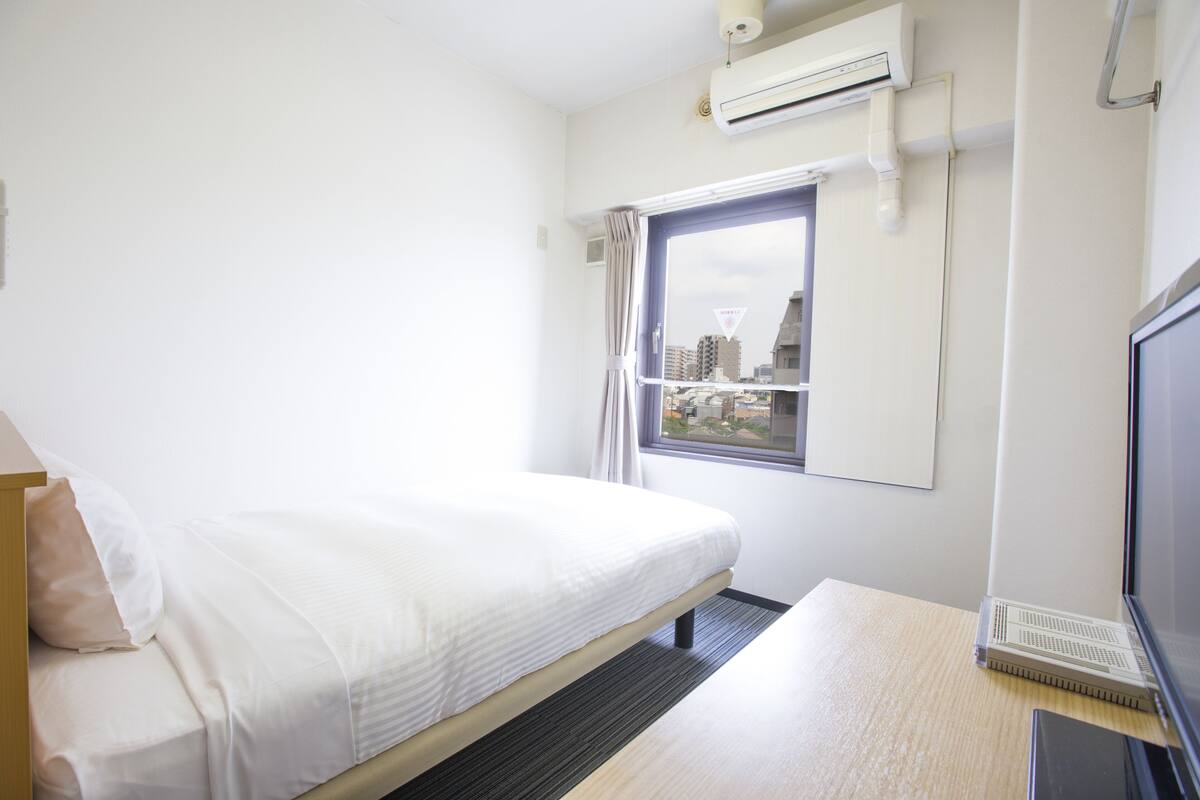 Une chambre d’hôtel équipée d’un lit, d’un bureau avec un ordinateur portable, d’une fenêtre donnant sur des immeubles et d’un appareil de climatisation.