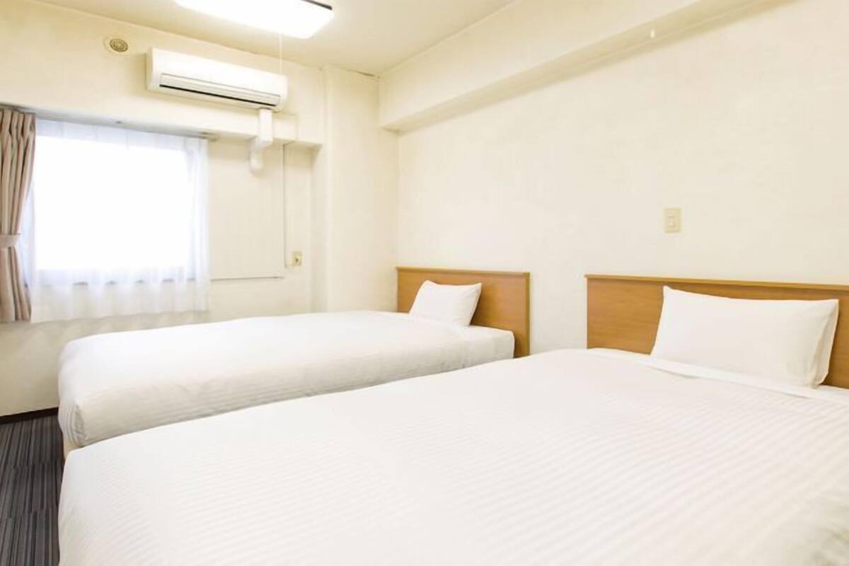 Une chambre d’hôtel avec deux lits simples, une fenêtre avec des rideaux, un appareil de climatisation et un mur uni.