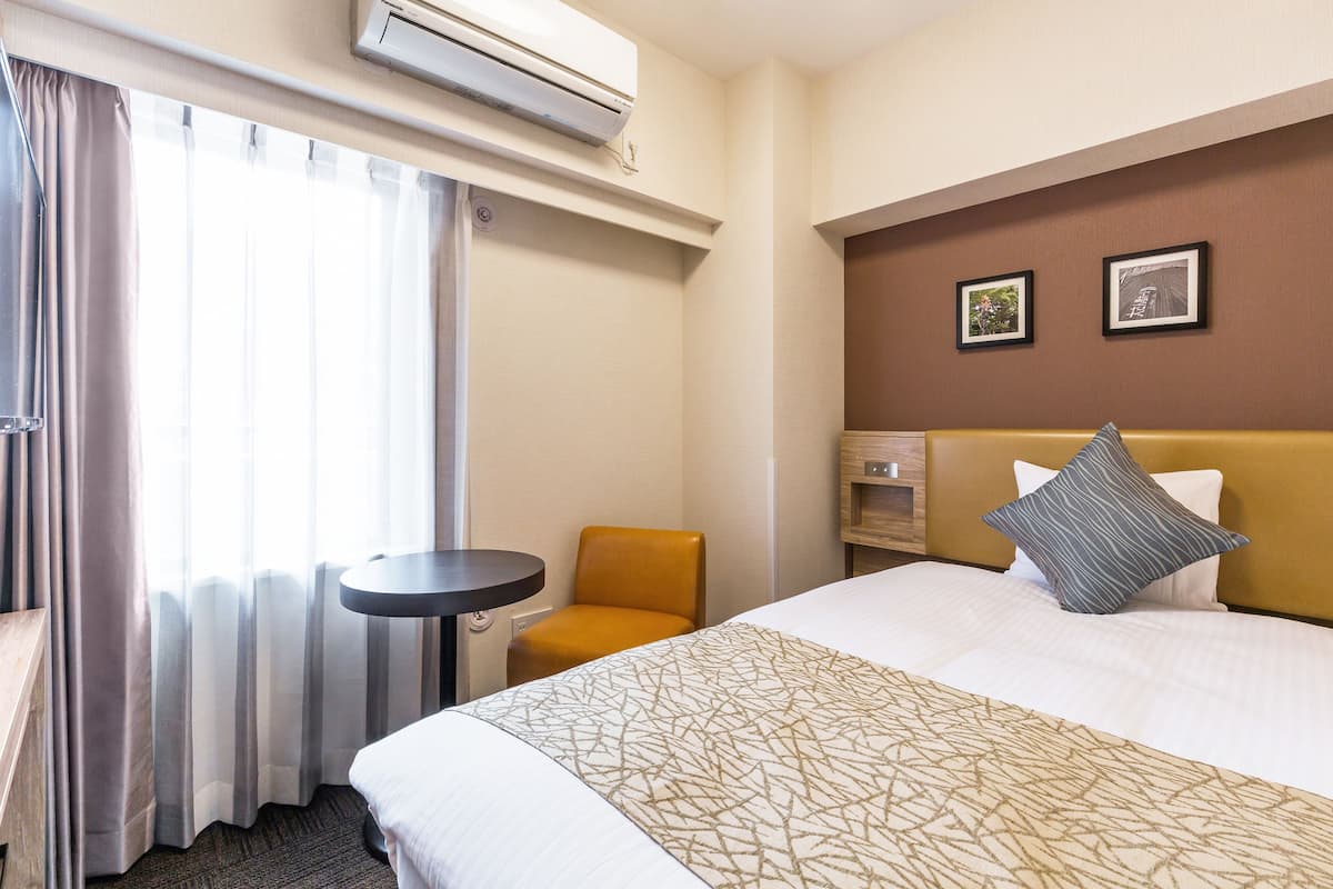 Ein Hotelzimmer mit einem Bett, einem kleinen Tisch, einem Sessel, einer Klimaanlage und zwei gerahmten Bildern an der Wand.