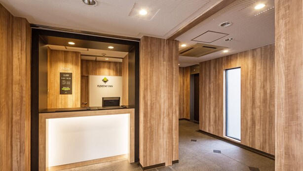 A reception area with a wooden reception desk, a sign indicating the name 'FLEXTAY INN', and a view of a hallway with a large window. - Flexstay Inn Sakuragicho (Yokohama)