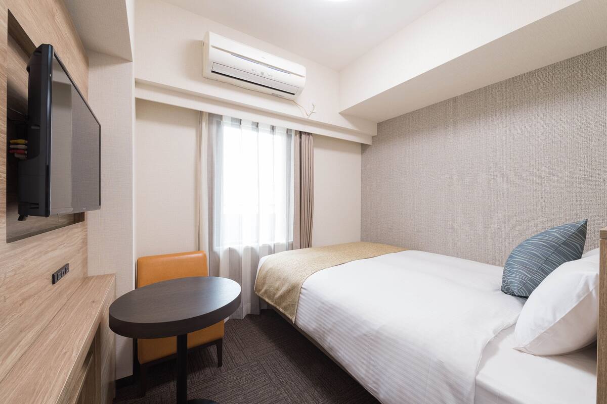 Ein Hotelzimmer mit Bett, Wandfernseher, kleinem Tisch und einem fenster mit Vorhängen.
