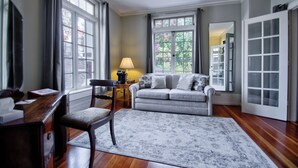 Una sala de estar con un escritorio de madera, un sofá gris, un tapete estampado y ventanas grandes con cortinas.
