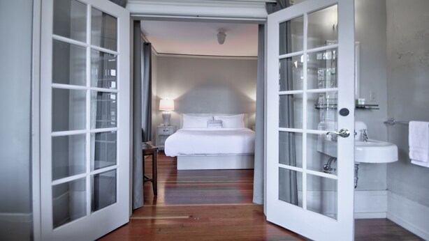 Habitación de hotel con cama, mesita de noche, lámpara y baño con lavabo y espejo.
