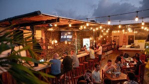 An outdoor bar area with wooden tables and chairs, a counter with a bartender, and a sign reading "CHEF HOBBY'S". - Che Playa Hostel & Bar (Playa del Carmen)