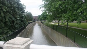 Un canal con una pasarela de hormigón a un lado y una zona arbolada y cubierta de césped al otro.