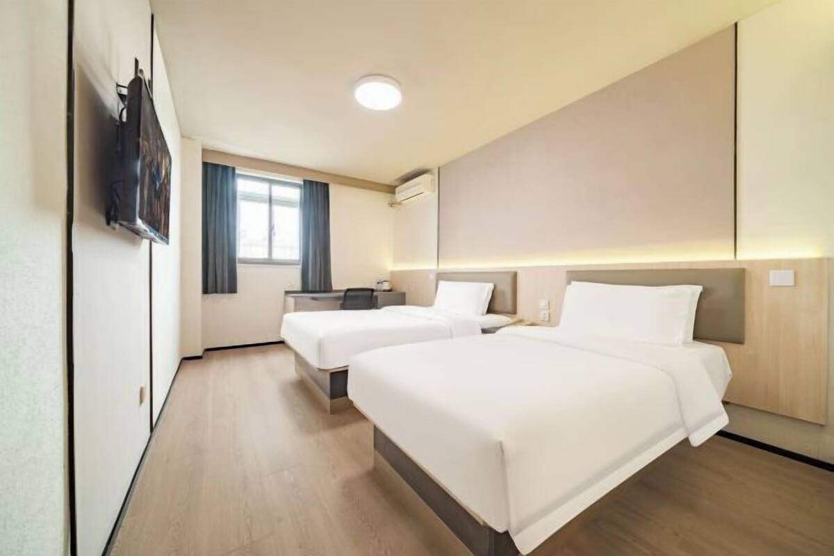 A hotel room with two beds, a desk, a flat-screen TV mounted on the wall, and a window with curtains.