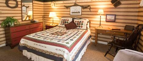 A log cabin-style room with a large bed, two bedside tables with lamps, a desk with a chair, a dresser, and a mirror.