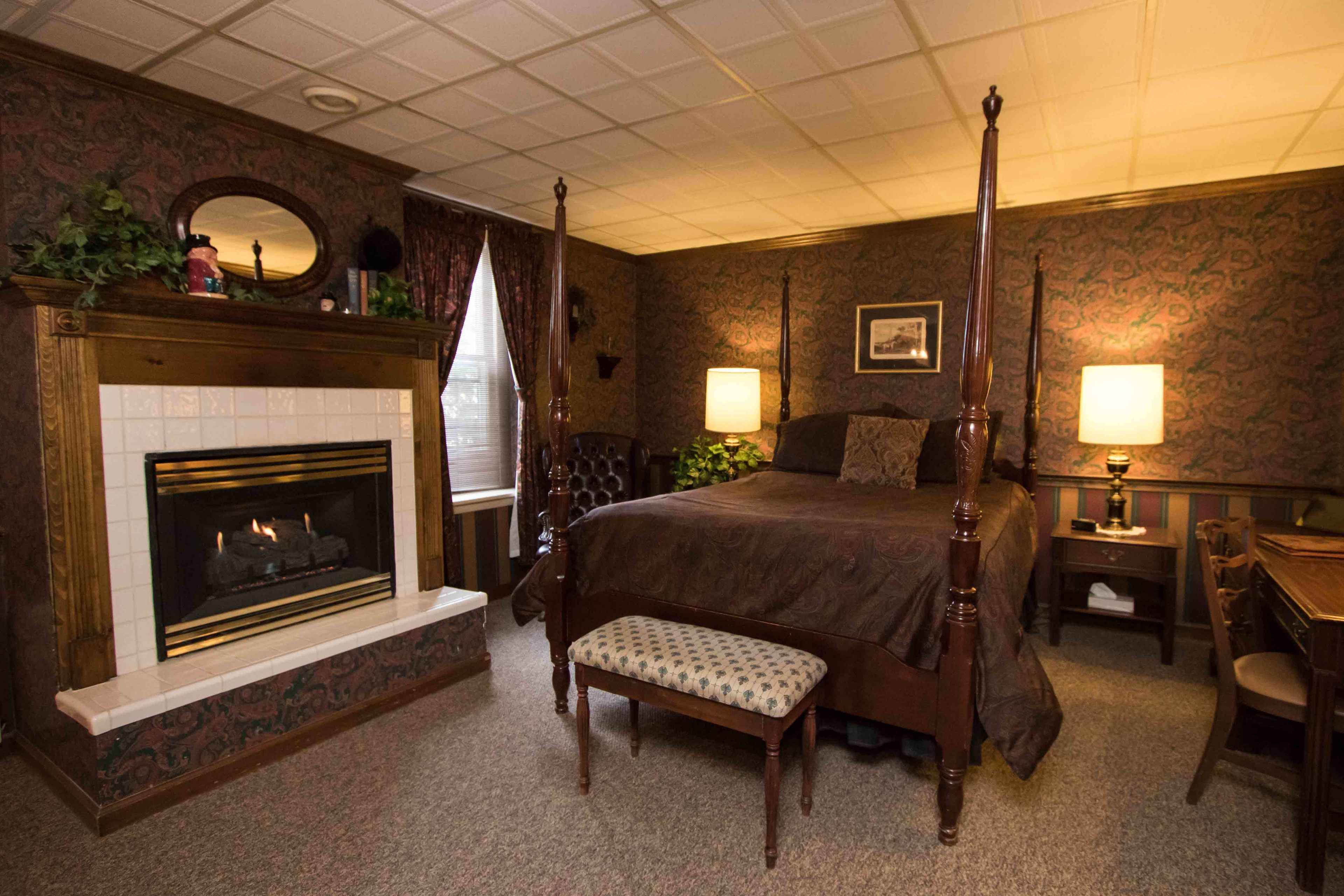 A four-poster bed, a fireplace, a bench, a desk, and two lamps in a hotel room.