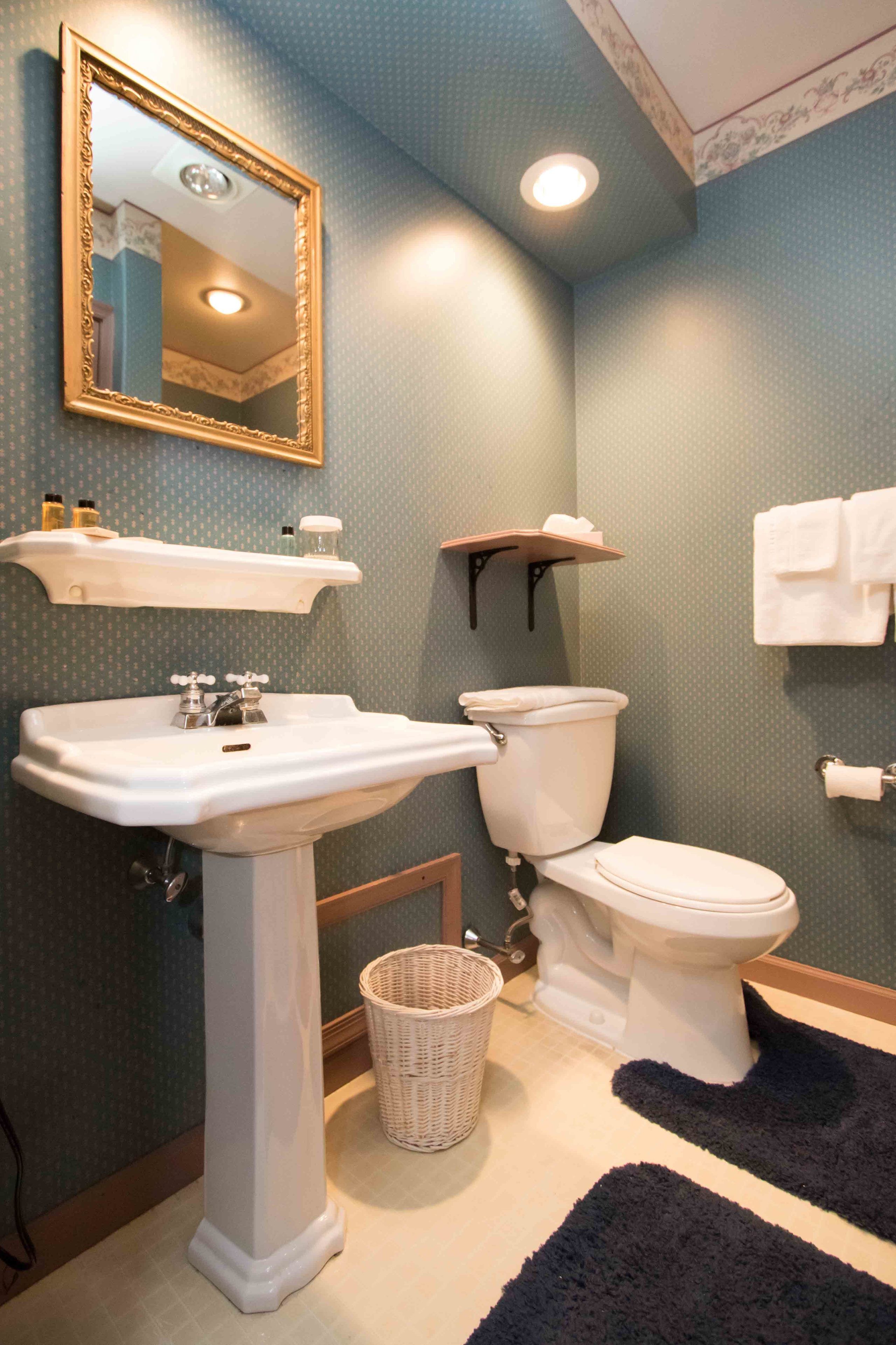 A bathroom with a pedestal sink, toilet, and a mirror with a decorative frame. There are shelves above the sink and a basket on the floor.
