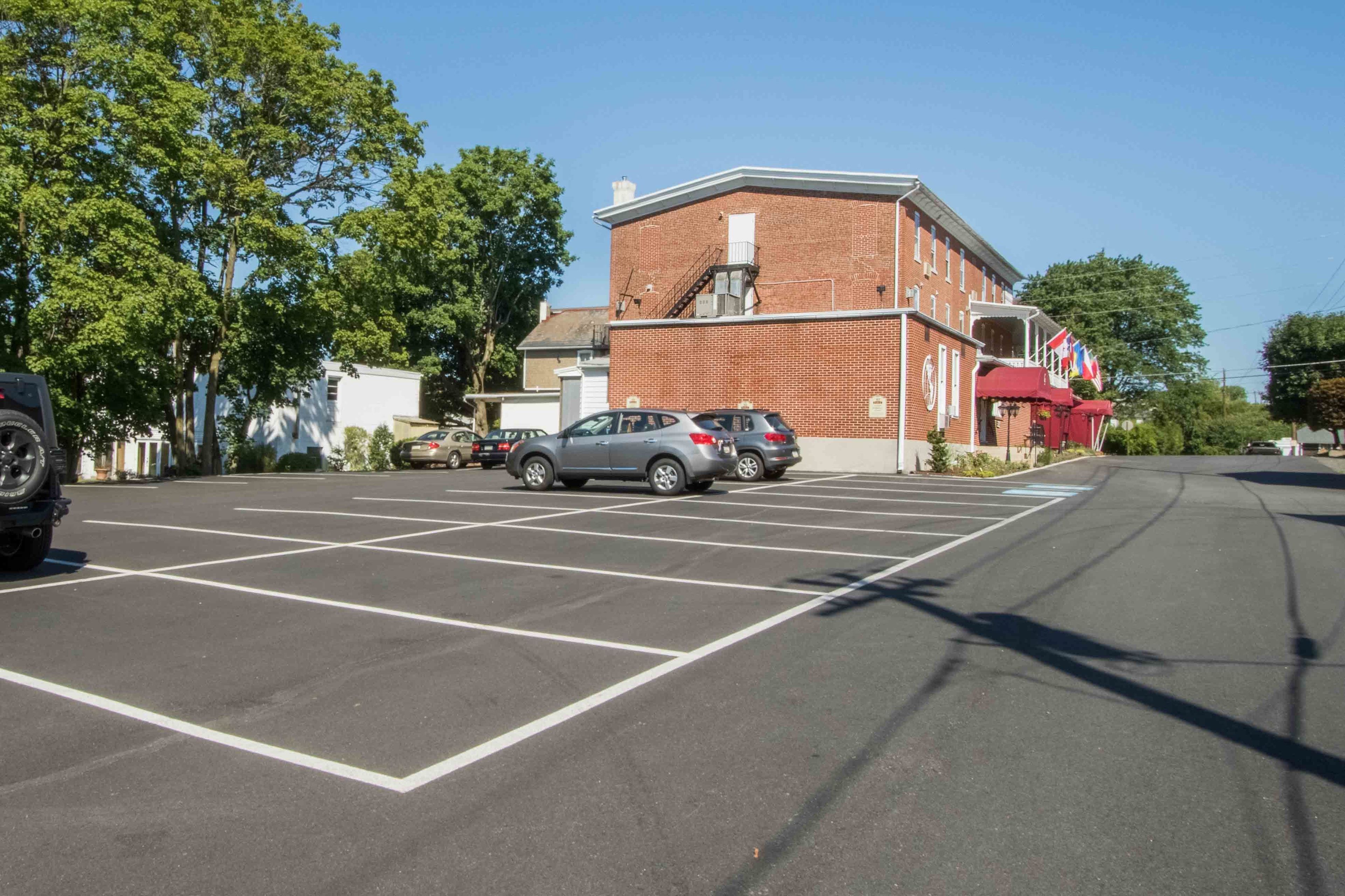 Un parking avec plusieurs voitures, un bâtiment en briques surmonté d’une marquise rouge, et des arbres en arrière-plan.
