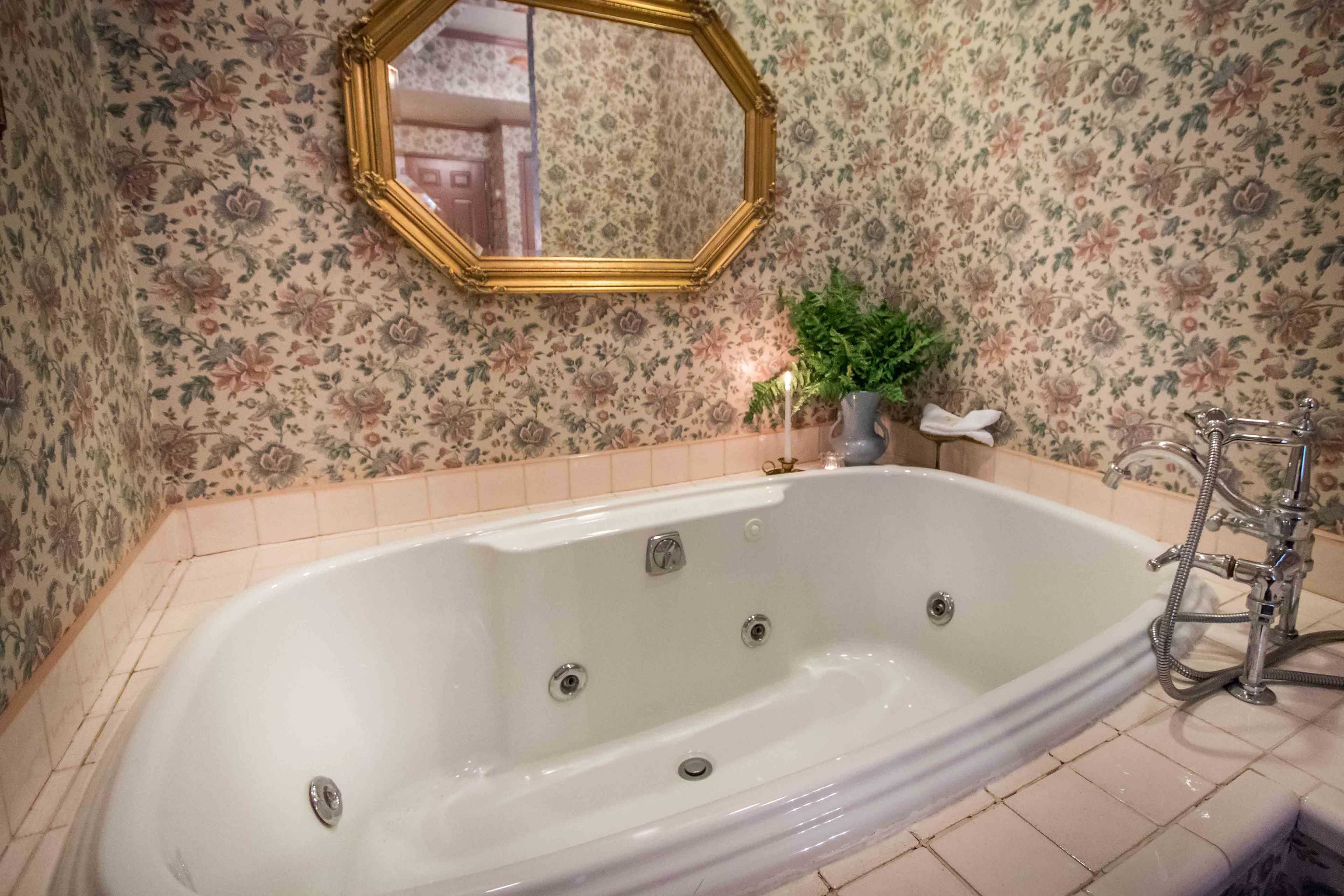 Un baño con una bañera grande, un espejo con marco dorado, papel tapiz floral y un perchero.