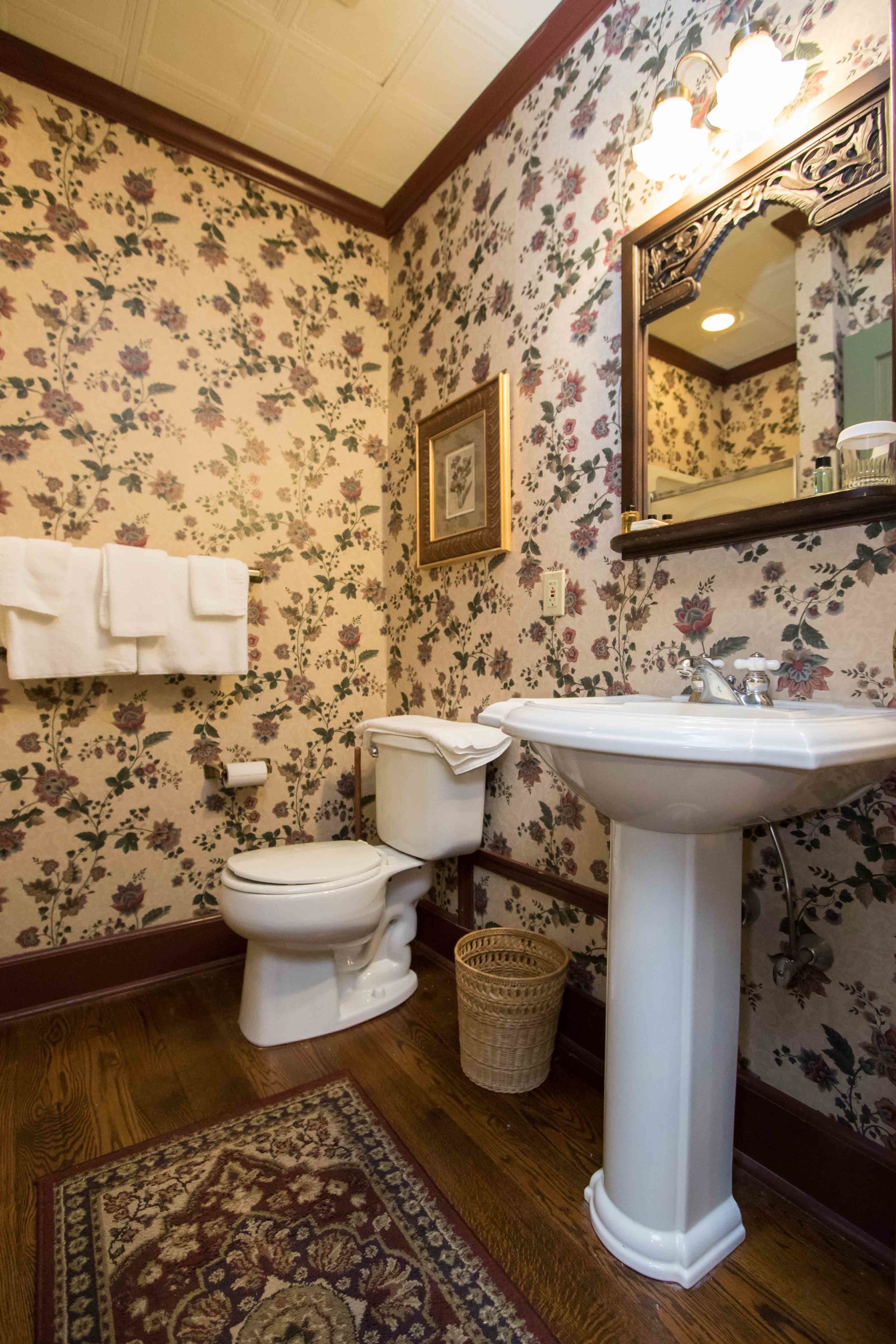 A bathroom with floral wallpaper, a white toilet, a pedestal sink, and a wicker basket.