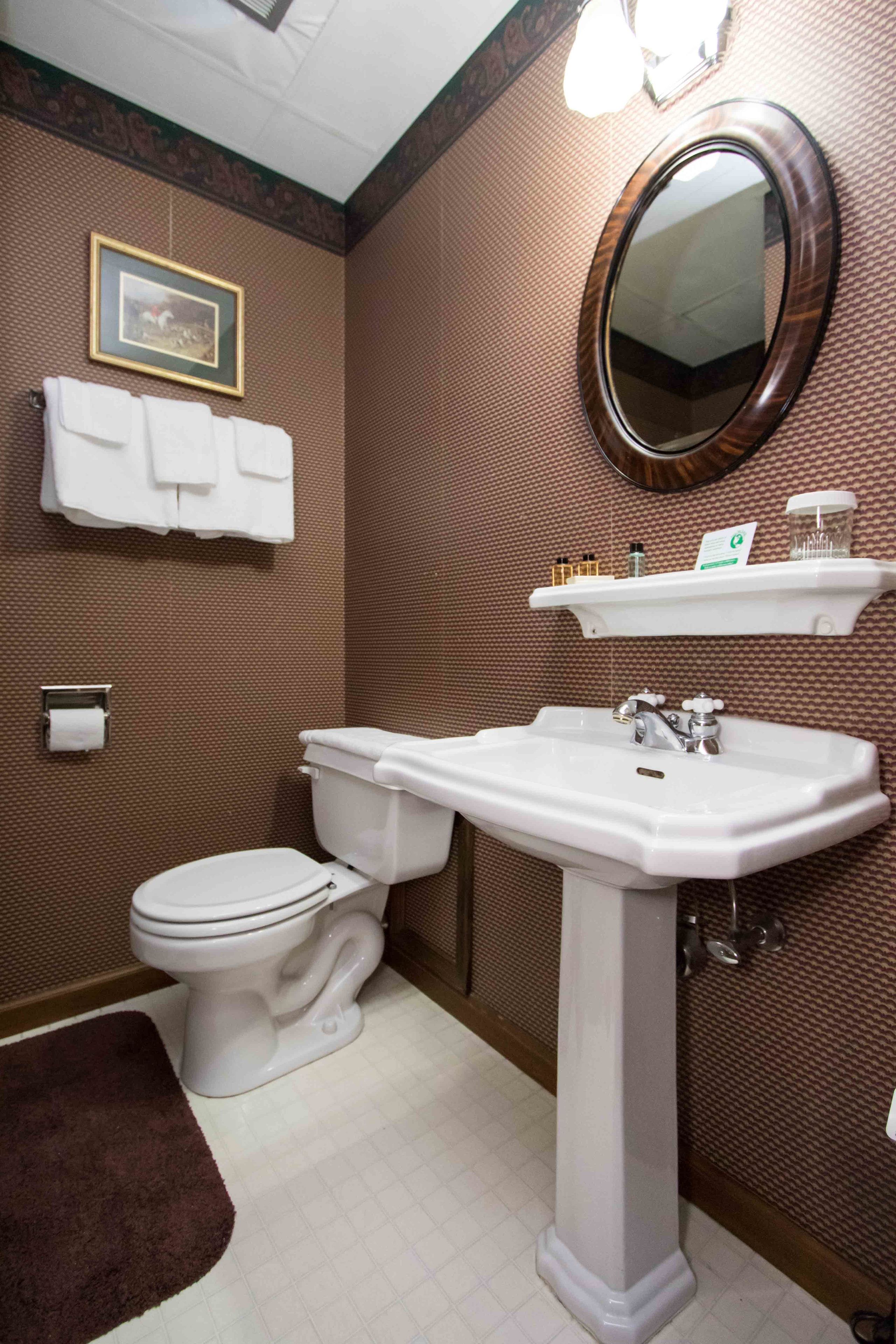 Une salle de bain comprenant des toilettes, un lavabo et un miroir. On y trouve un porte-serviettes avec des serviettes, un tableau accroché au mur et une étagère avec des articles de toilette.