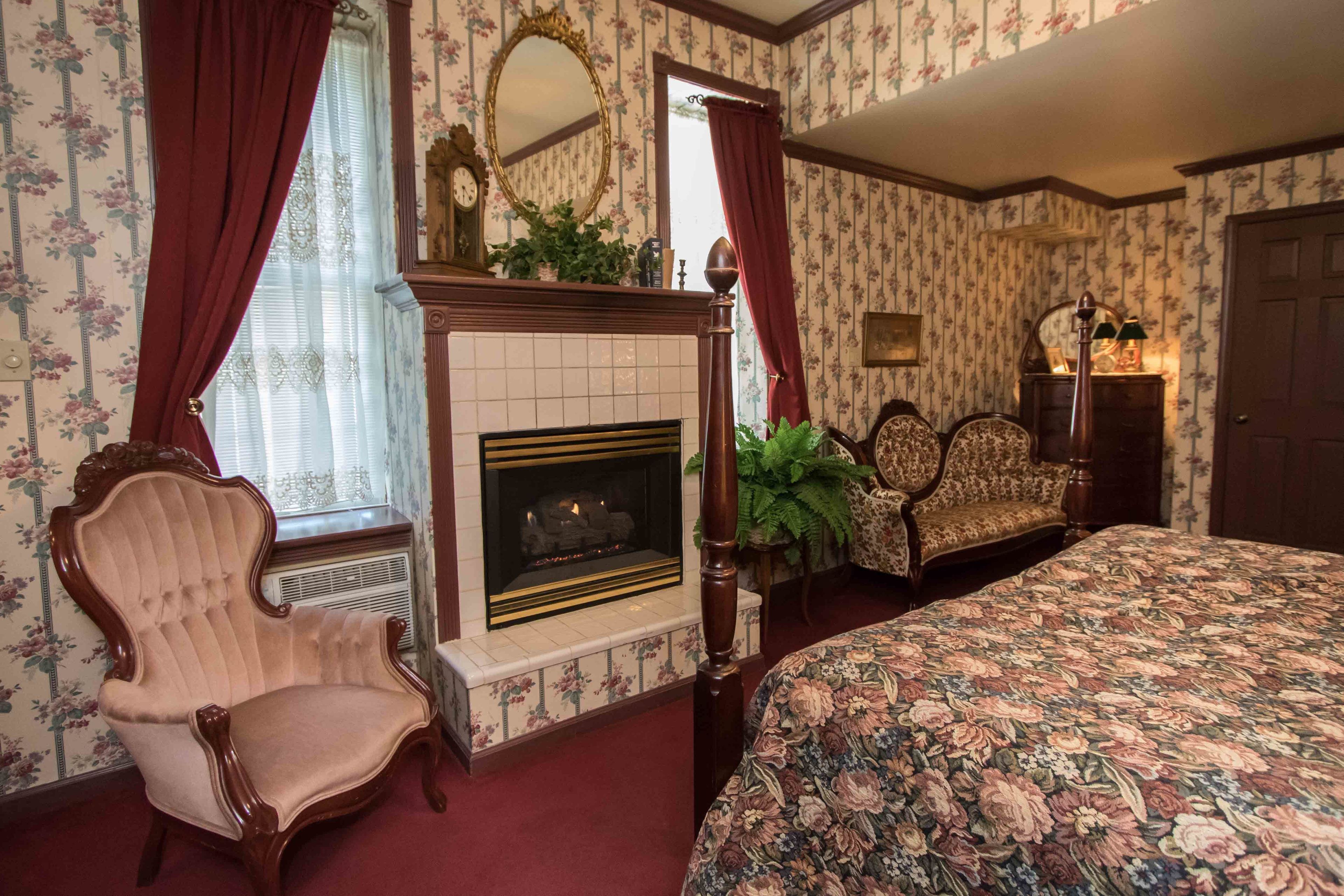 Un dormitorio con colcha estampada de flores, chimenea, sillón antiguo y cómoda de madera.