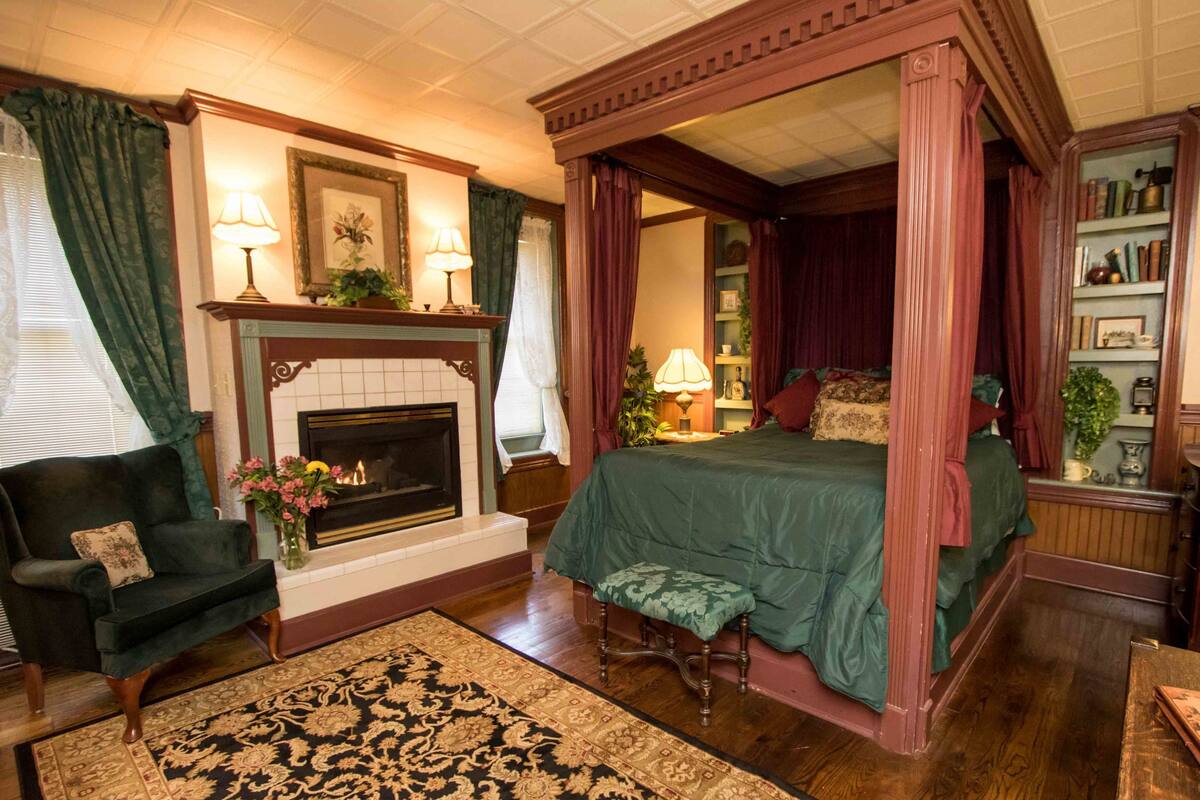 A four-poster canopy bed with a green bedspread, a fireplace with a mantel, a dark green armchair, and a patterned rug.