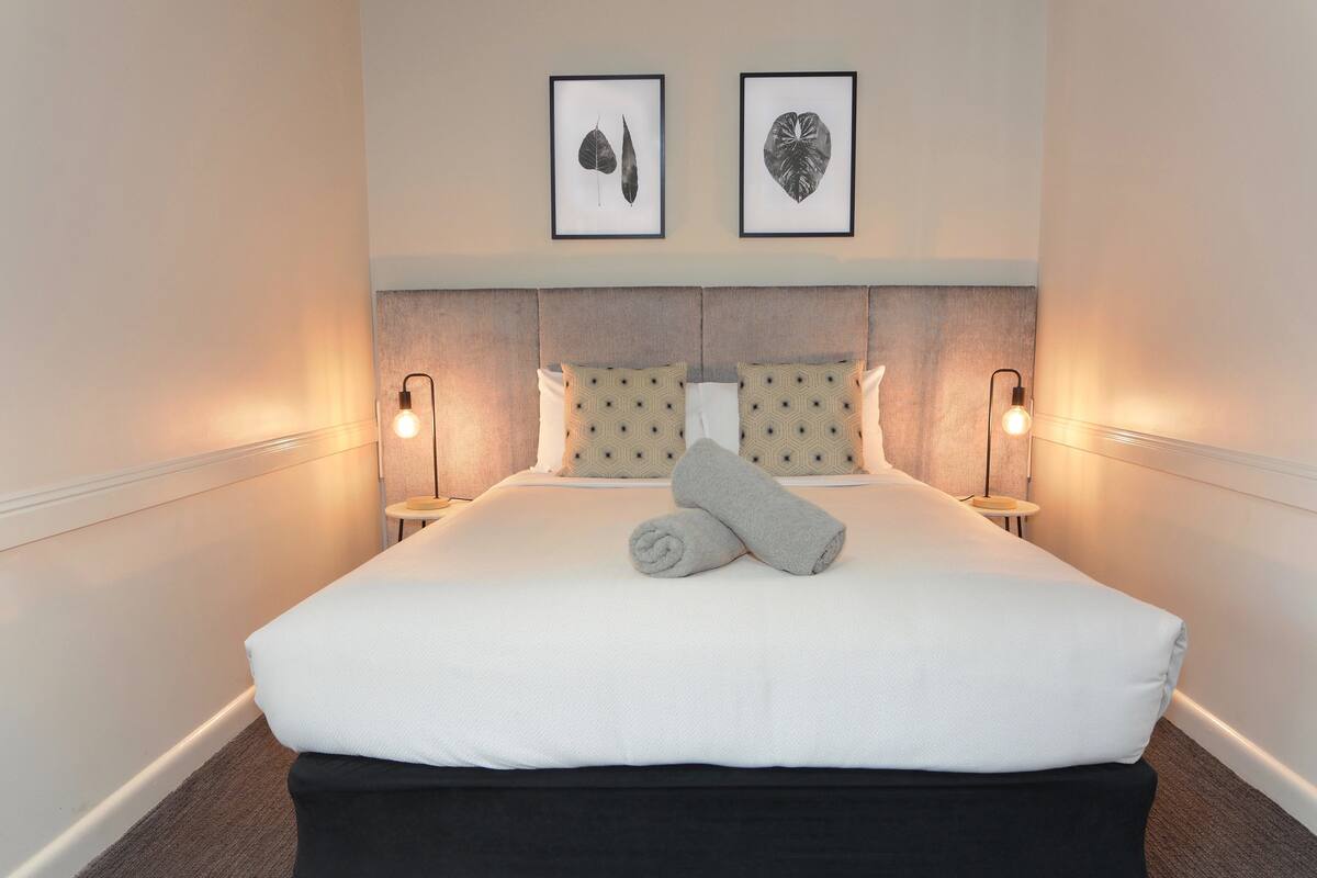 A neatly made bed with a white comforter and two rolled towels, a headboard with a textured design, and two framed leaf artworks on the wall.