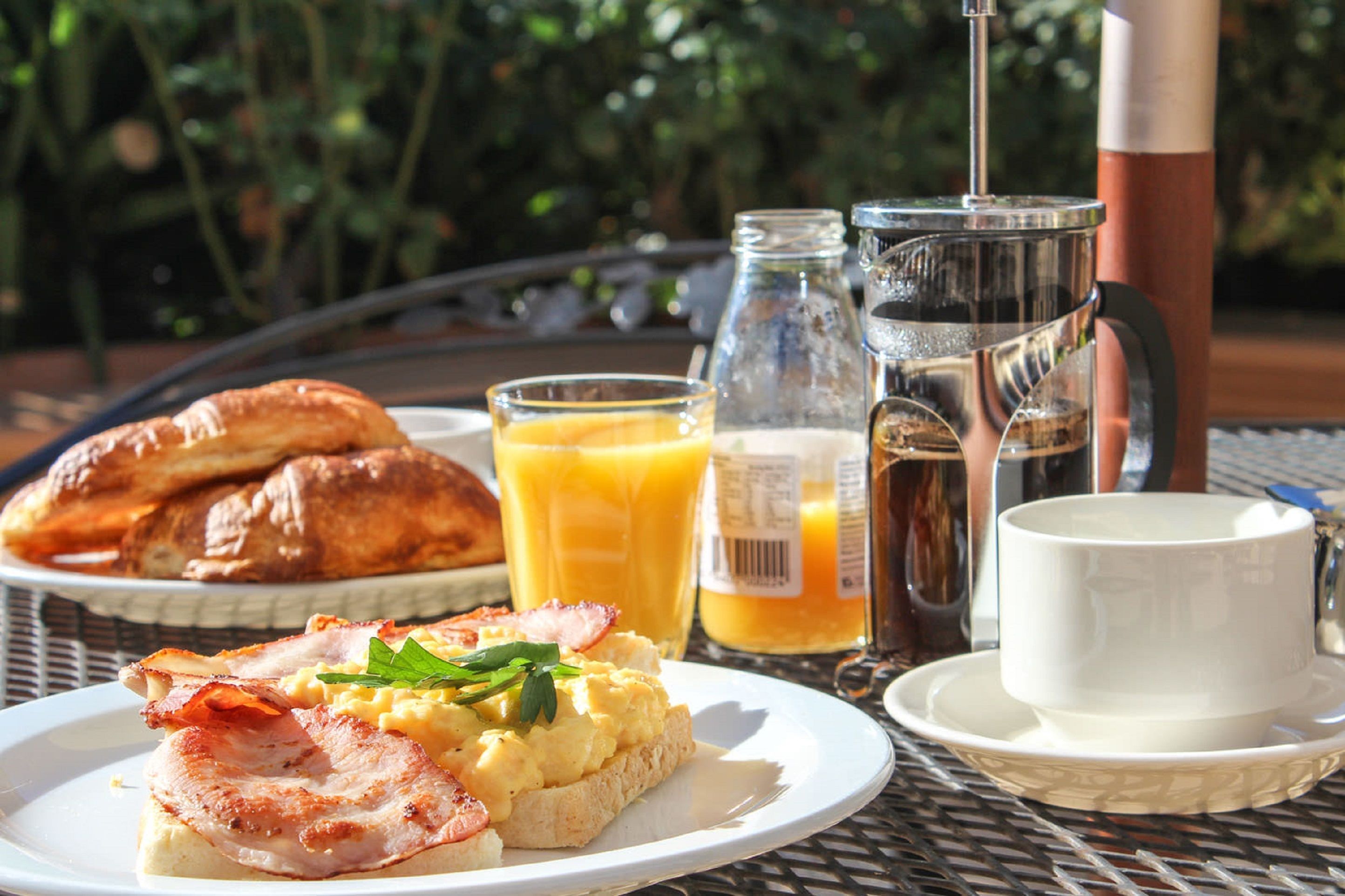 Een ontbijtsetting met croissants, roerei, en spek op een bord, een glas sinaasappelsap, een koffiepot en een kopje op een tafel buiten.