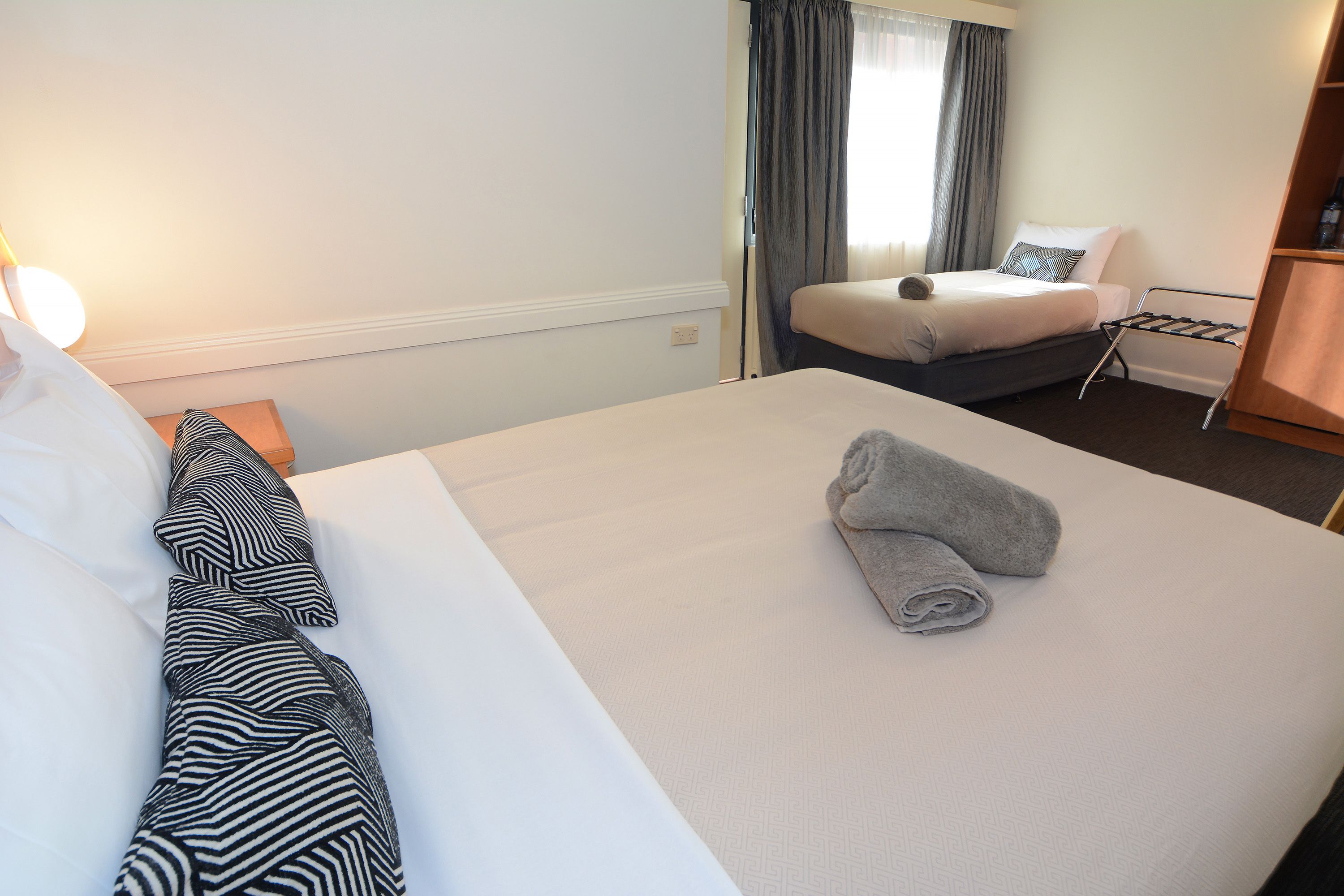 A hotel room with a large bed, a bedside table, a lamp, a window with curtains, and a small seating area with a chair and a table.