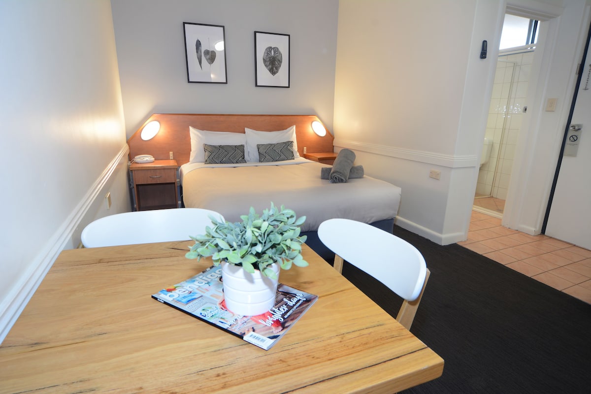 A neatly arranged bedroom with a bed, bedside tables, a nightstand, a bedside lamp, a potted plant, and a magazine on a wooden table.