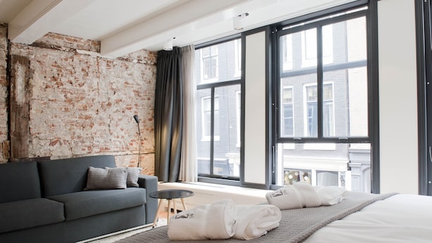 Una habitación de hotel moderna con pared de ladrillo, un sofá gris, cama con ropa de cama blanca y ventanas amplias que ofrecen vistas a la ciudad.
