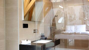 A modern bathroom with a glass shower enclosure, a black countertop sink, and a visible bed in the adjacent room.