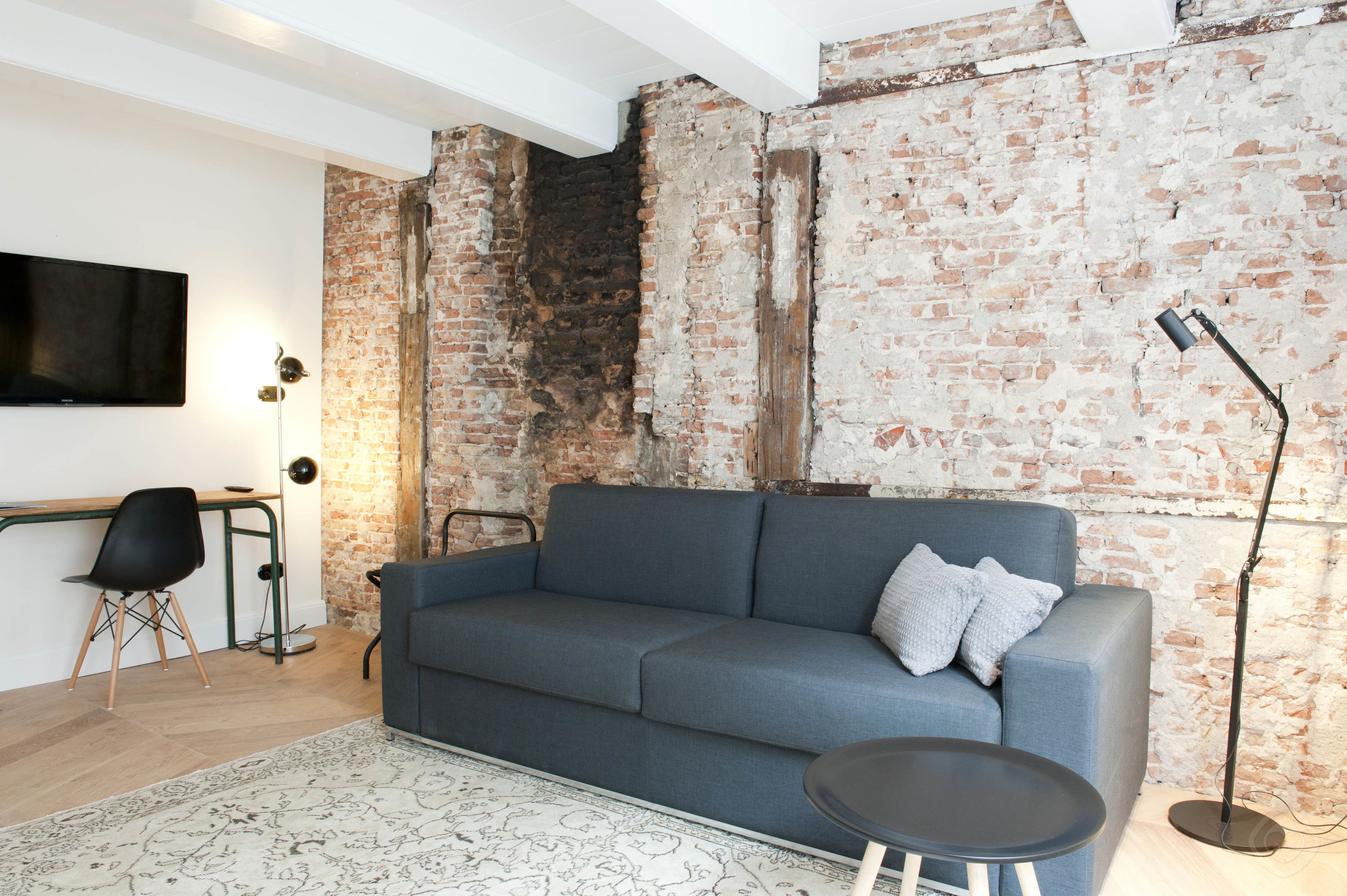 A modern living room with a grey sofa, a small round table, a TV mounted on the wall, and a brick wall.