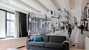 Una sala moderna con un ventanal grande, un sofá gris con cojines azules, un taburete redondo de madera y un mural de la calle en la pared.