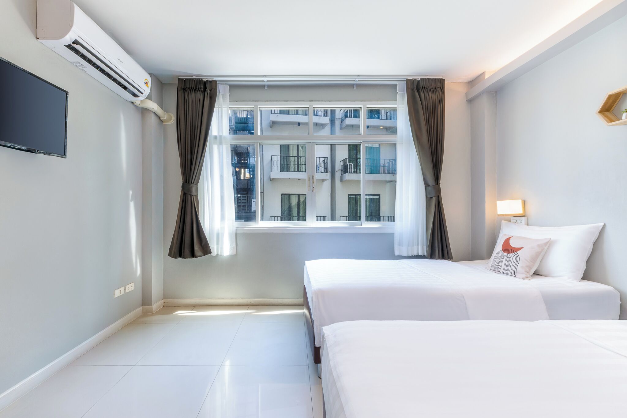 A hotel room with two beds, a flat-screen TV, an air conditioner, a window with curtains, and a view of a building outside.